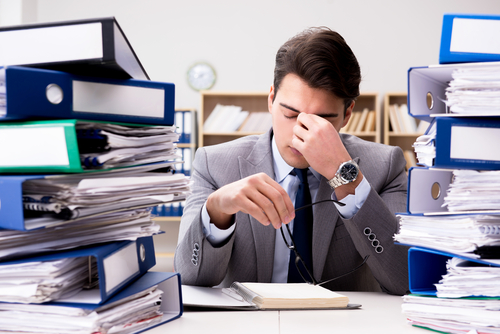 A stressed man at a desk between two tall stacks of binders and paperwork, illustrating the article on a fiduciary's withholding and reporting duties on distributions to a beneficiary in Germany