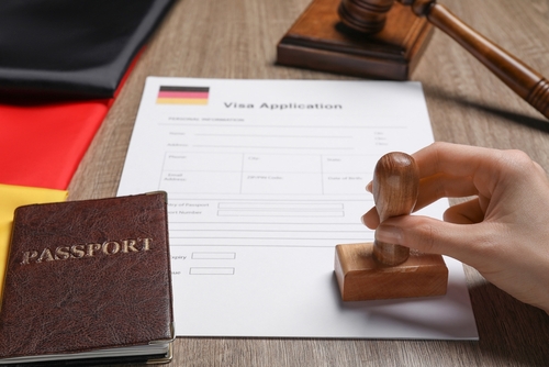 A hand stamping a German visa application form next to a passport, a German flag, and a gavel, illustrating the article on relocating to Germany while holding a U.S. trust